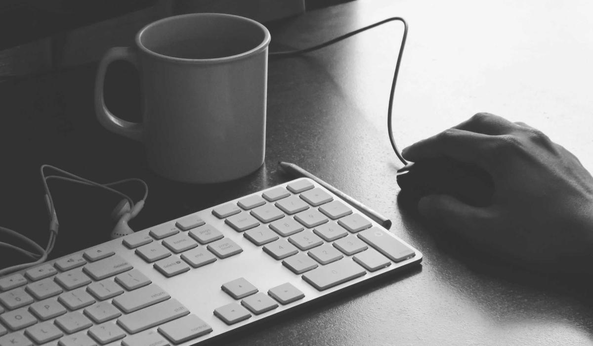 A hand on a computer mouse next to a keyboard and a cup of coffee